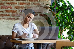 Beautiful young girl working at a coffee shop with a laptop and taking notes. Female freelancer connecting to internet via