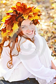 Beautiful young girl in a wreath from yellow leaves, in white clothes, sitting on fall leaves on a Sunny day and laughing