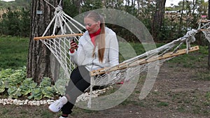 Beautiful young girl writes a message to use a smartphone sitting in hammock smiling