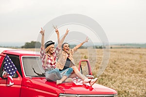 beautiful young girlfriends sitting on car hood with raised hands and showing