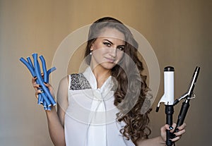 Beautiful young hairstulist with brunette curly hair holding blue rollers and iron curler in her hands trying to make a choice whi