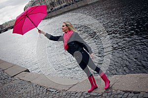 Beautiful young and happy blond woman in a bright pink scarf, rubber boots and umbrella walking in a rainy city.