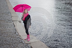 Beautiful young and happy blond woman in a bright pink scarf, rubber boots and umbrella walking in a rainy city.