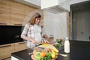 Beautiful pregnant woman chopping yellow bell pepper on a cutting board, preparing healthy meal in the kitchen island