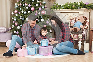 Beautiful young happy family caucasian mother father and child in plaid shirt and jeans sitting near christmas tree and fireplace