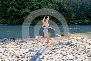 Beautiful young happy fit woman running on sandy beach with dog enjoying nature.