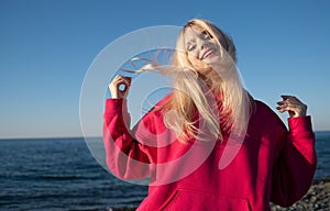 A beautiful, young, happy and smiling blonde girl in a pink hoodie rejoices against the backdrop of the sea and sky. The
