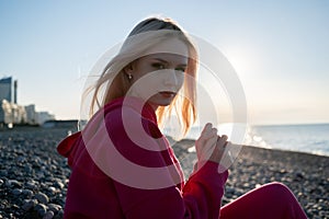 A beautiful, young, happy and smiling blonde girl in a pink hoodie rejoices against the backdrop of the sea and sky. The