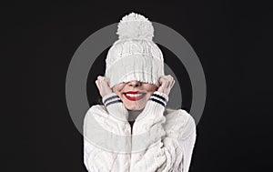 Beautiful young happy smiling girl hiding her eyes under trendy white big loop knitted beanie hat. Model wearing winter sweater.