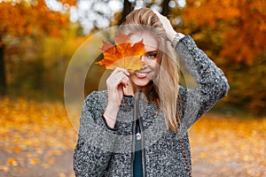 Beautiful young happy woman in a fashionable gray coat holding a bright yellow autumn leaf near the face in the park. Joyful girl