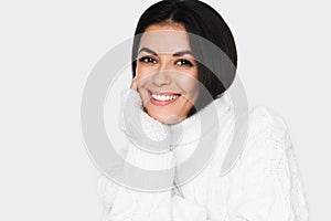Beautiful young happy woman looking pensive and happy in white sweater on light grey background.