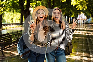 Beautiful young happy women friends walking outdoors with backpacks showing peace gesture.