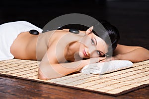 Beautiful, young and healthy woman on bamboo mat in spa salon is having hot stone massage.