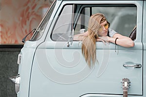 A beautiful young hippie woman in yellow sunglasses looks out the window of a blue vintage minivan. Make love, not war