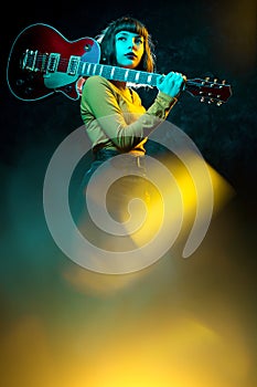 Beautiful young hipster woman with curly hair with red guitar in neon lights. Rock musician is playing electrical guitar