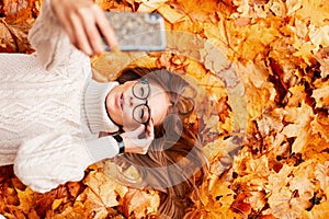 Beautiful young hipster woman is lying in the park on orange leaves in the park and makes a selfie on a modern smartphone.