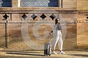 Beautiful young Hispanic woman, brown-haired, wearing a striped sweater, linen pants and sunglasses, walking with her suitcase at