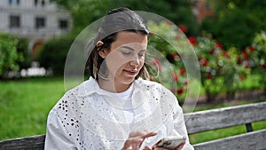 Beautiful young hispanic woman smiling happy using smartphone sitting on a bench at the park