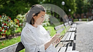 Beautiful young hispanic woman using smartphone sitting on a bench at the park