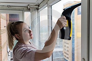 Beautiful young housewife in working clothes washing windows by professional tools at home.