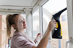 Beautiful young housewife in working clothes washing windows by professional tools at home.