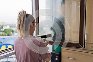Beautiful young housewife in working clothes washing windows by professional tools at home.