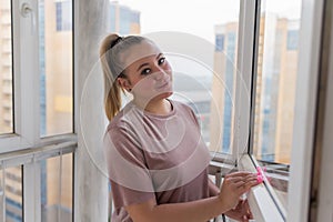 Beautiful young housewife in working clothes washing windows by professional tools at home.
