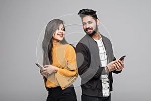 Beautiful young indian couple holding mobile phones and standing back to back against grey background