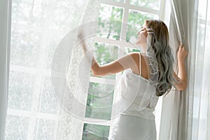 Beautiful young joyful Asian woman with long gray hair wake up in morning opening window white curtains and looking out of window