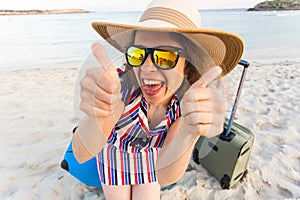 Beautiful young lady with a blue suitcase showing thumbs up gesture on the beach. People, travel, vacation and summer