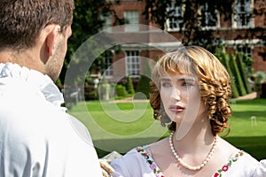 Beautiful young lady dressed in period costume confronting her husband or lover with stately home in background.