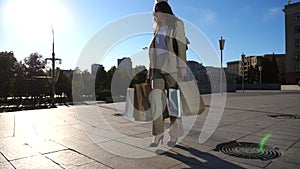 Beautiful young lady holds shopping bags going at city square after purchases. Fashionable woman carries paper packets