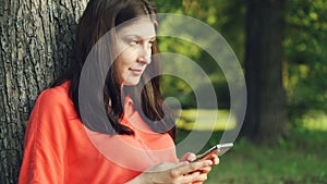 Beautiful young lady is using smartphone resting in park under tree and enjoying modern technology and summer nature