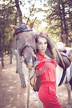 Beautiful young lady wearing red dress riding a horse at sunny summer day. Brunette with long curly hair with flowers on her head