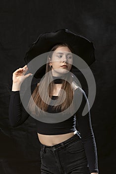 Beautiful young lady wears black clothes and wide-brimmed hat. Portrait of young model on black background. Vertical frame