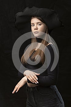 Beautiful young lady wears black clothes and wide-brimmed hat. Portrait of young modern aristocrat on black background. Vertical