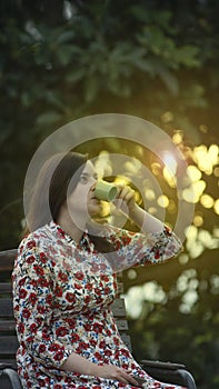 Beautiful young latin woman in floral design dress drinking a cup of coffee in the garden at sunset with the sun`s rays passing