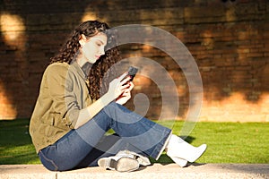 Beautiful young latina woman with curly brown hair and dressed in casual clothes sitting on a grey wall checking her mobile phone