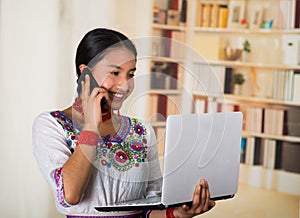 Beautiful young lawyer wearing traditional andean blouse and red necklace, holding laptop talking on phone smiling