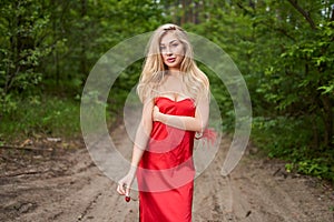 Beautiful young long-haired blonde in a red dress posing on the road in a summer park with a ripe strawberry fruit in her hand.