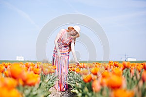 Beautiful young long red hair woman wearing in striped dress and straw hat standing on colorful flower tulip field in Holland