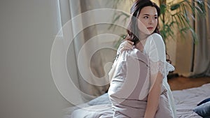 Beautiful young millennial brunette bride posing for camera, sitting on mattress in luxurious white peignoir and