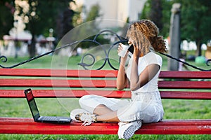 Beautiful Young Mixed Race Woman Working With Laptop And Using Phone. Sitting On The Bench In Park, Outdoor Portrait , Wears