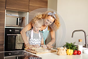 Beautiful young mom and her cute little daughter rolling out dough at home.
