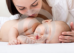 Beautiful young mom looking at her sleeping little baby and smiling. Child lying in bed at home