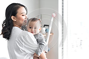 Beautiful young mom is playing with her cute baby and smiling while standing near the window at home