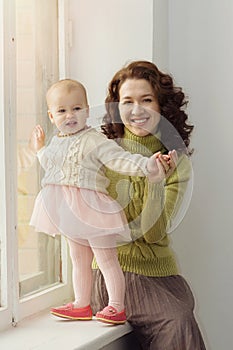 A beautiful young mother with curly dark hair in a cozy knitted sweater sits at the window with a little girl in her