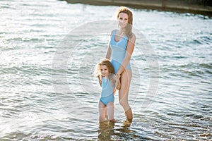 Beautiful young mother and daughter having fun resting on the sea. They stand in the water in the same swimsuit and smile
