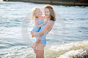 Beautiful young mother and daughter having fun resting on the sea. They stand in the water in the same swimsuit and smile. mother