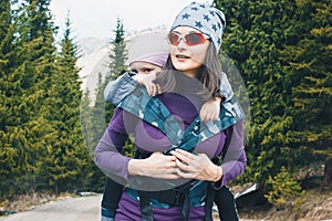 Young mother with her toddler daughter on back in ergonomic baby carrier outside in nature. Babywearing concept